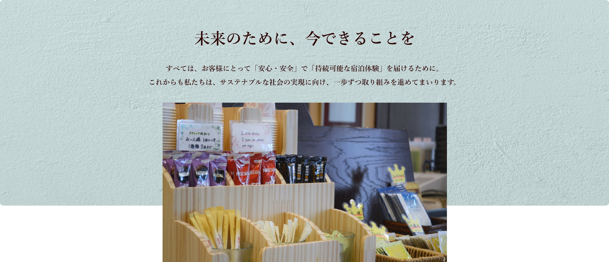 「未来のために、今できることを」というメッセージとともに、ホテルのアメニティコーナーが写っている画像。木製の棚に、スティックタイプのコーヒーやティーバッグ、使い捨てスプーンなどが整然と並べられており、「1人1本まで」などの案内が掲示されている。サステナブルな宿泊体験の実現を目指す取り組みを表している。